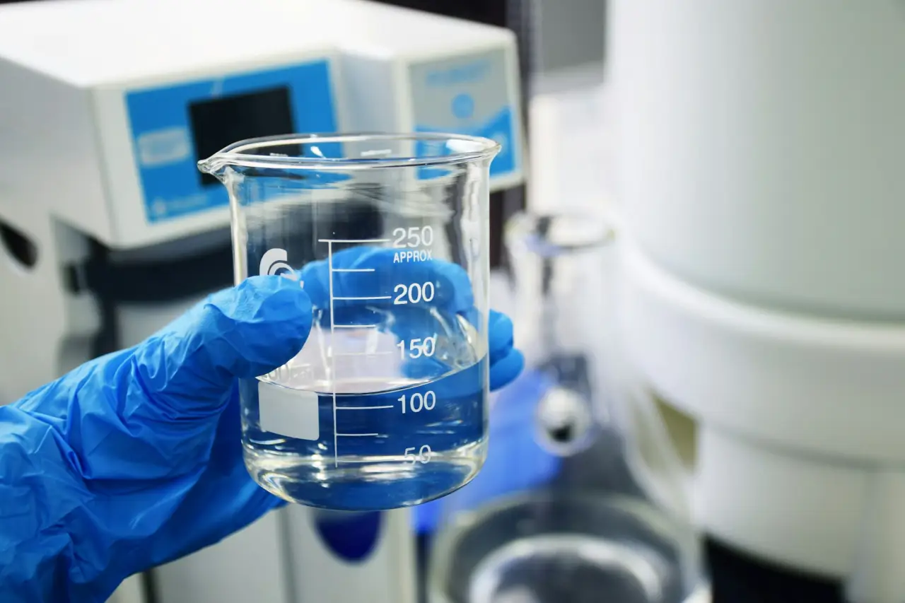 Gloved hand holding a glass beaker of clear liquid in a laboratory